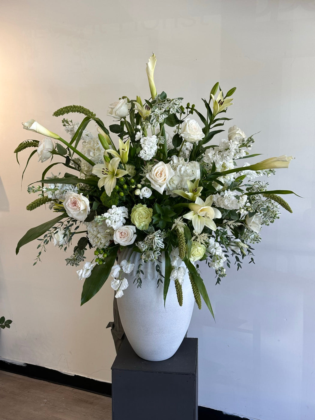 Tall white urn arrangement by Novapetal Flower Lab on a plinth, with white roses, lilies, stock and hydrangea, the kind of piece we build for a lobby or entrance