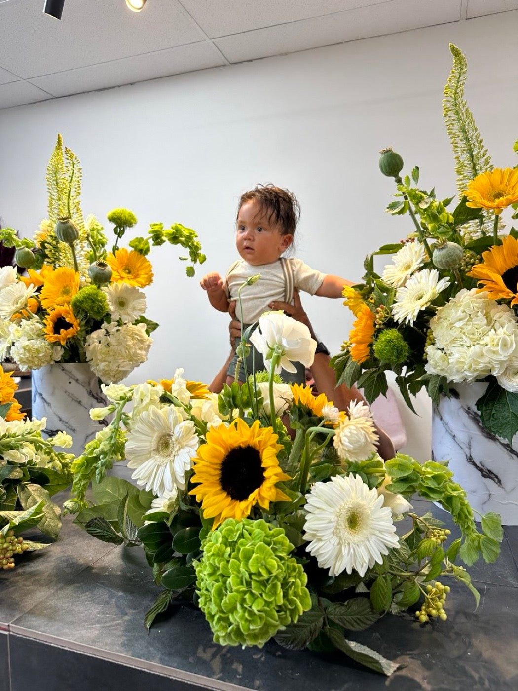 The founders son standing on the studio bench in Phoenix among arrangements of sunflowers, white hydrangea, gerbera and green viburnum