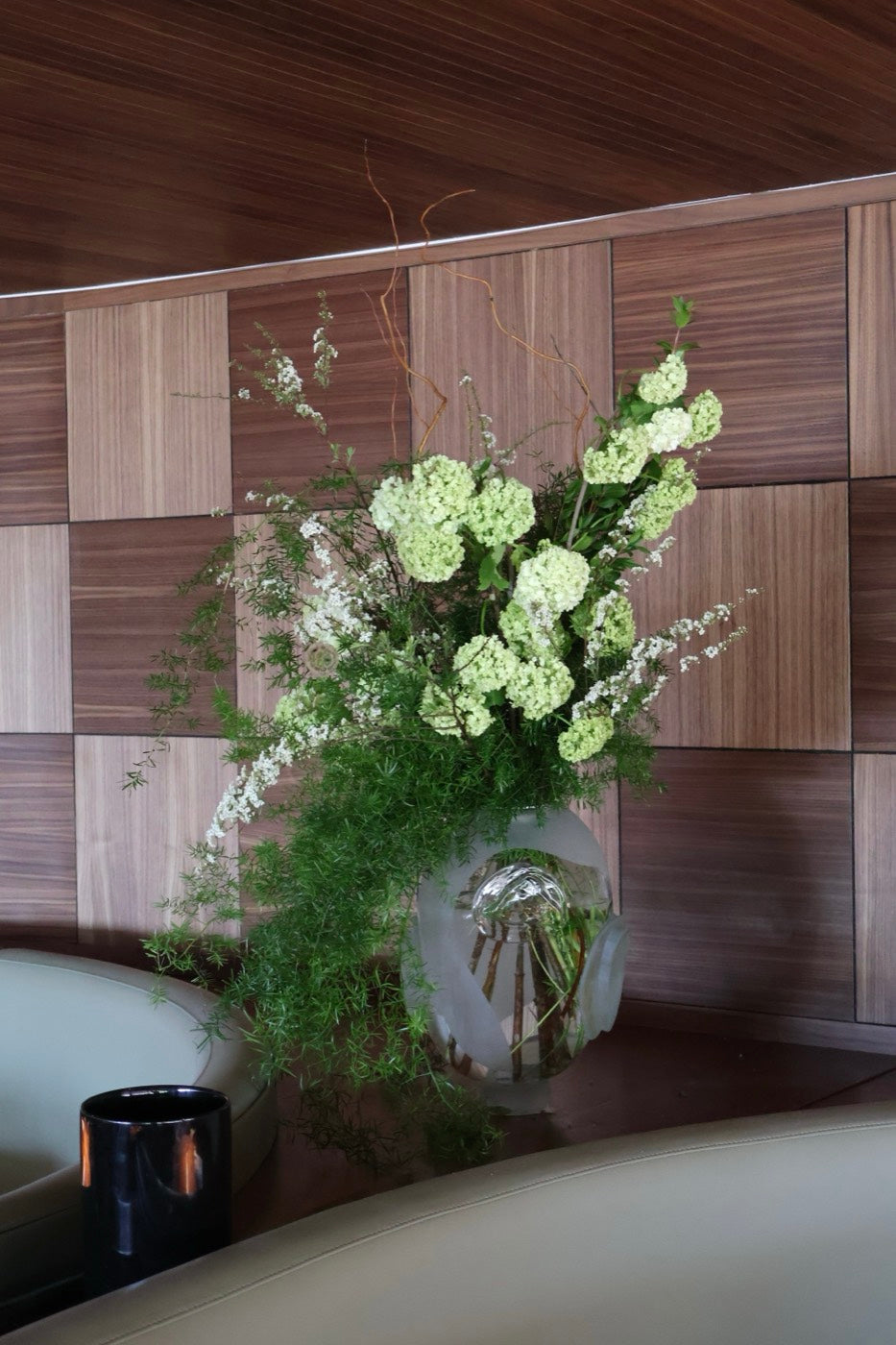 Tall green and white arrangement by Novapetal Flower Lab with viburnum, spirea and asparagus fern in a frosted glass vase, against the walnut panelling of a restaurant interior