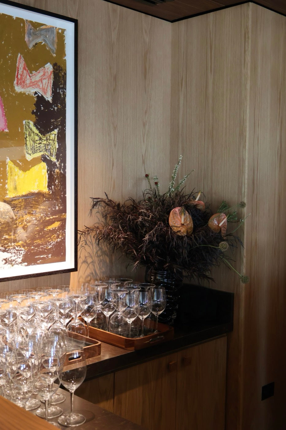 Dried protea and fern arrangement by Novapetal Flower Lab on a restaurant service bar beside stacked glassware, under a framed painting