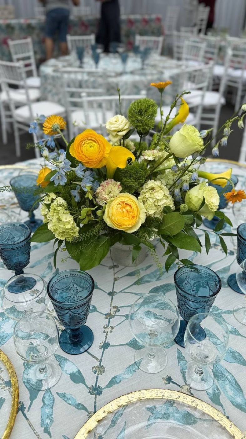 Event table styled by Novapetal Flower Lab with a low centerpiece of yellow ranunculus, garden roses, calla lilies and hydrangea, set with blue glassware and gold chargers under a marquee