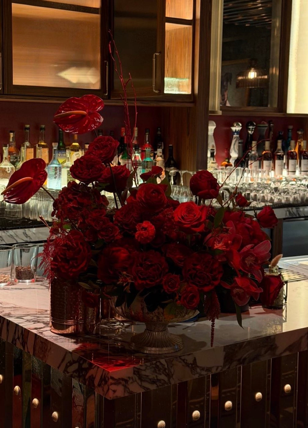 Red garden roses, ranunculus, anthurium and cymbidium orchids by Novapetal Flower Lab on the marble bar, the back bar and glassware behind