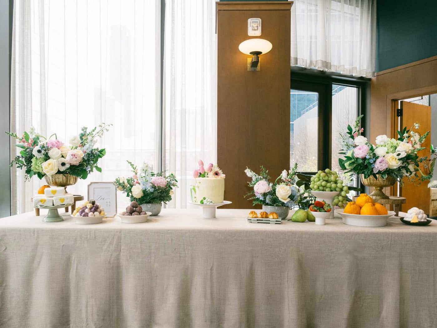 Celebration table styled by Novapetal Flower Lab at a Phoenix hotel, with two footed arrangements of white roses, pink peonies, anemones and delphinium along a linen-covered table set with desserts and fruit