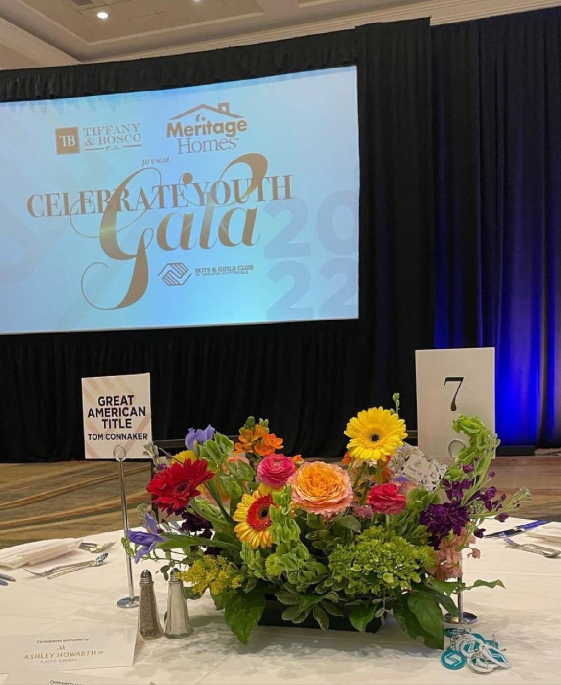 Long low centerpiece by Novapetal Flower Lab on a banquet table at the Celebrate Youth Gala in Phoenix, with gerbera, garden roses, hydrangea and sunflowers in front of the stage screen