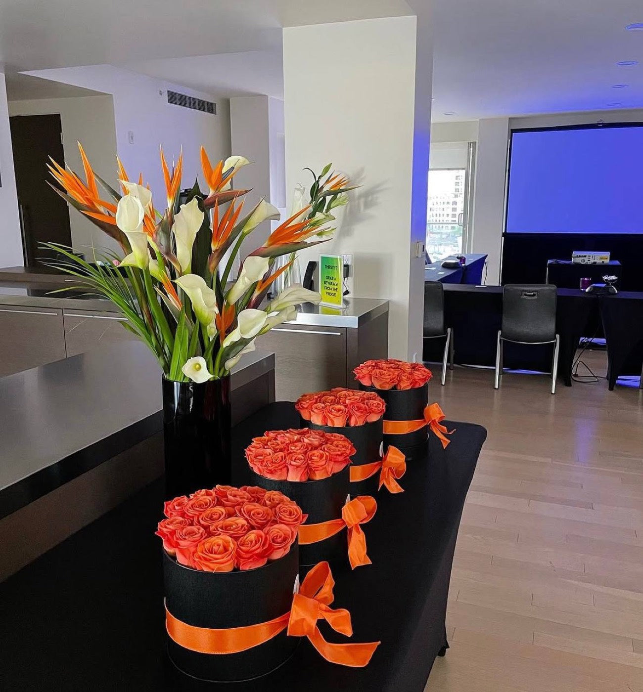 Row of black hat boxes of orange roses tied with orange ribbon by Novapetal Flower Lab, with a tall arrangement of birds of paradise and calla lilies, staged for a corporate event