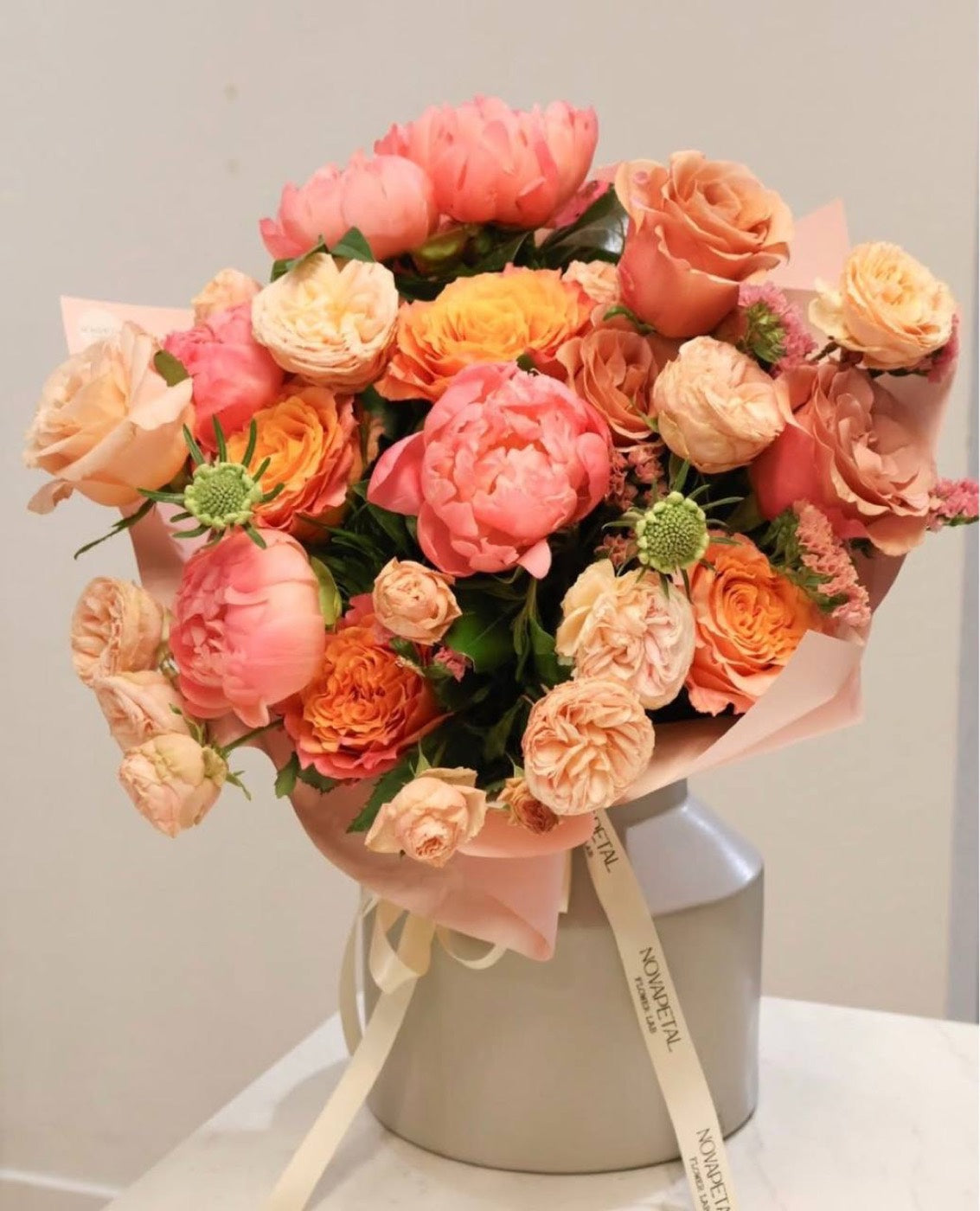 Peach and coral bouquet by Novapetal Flower Lab with garden roses, spray roses, a coral charm peony and craspedia, wrapped in blush paper with a Novapetal ribbon