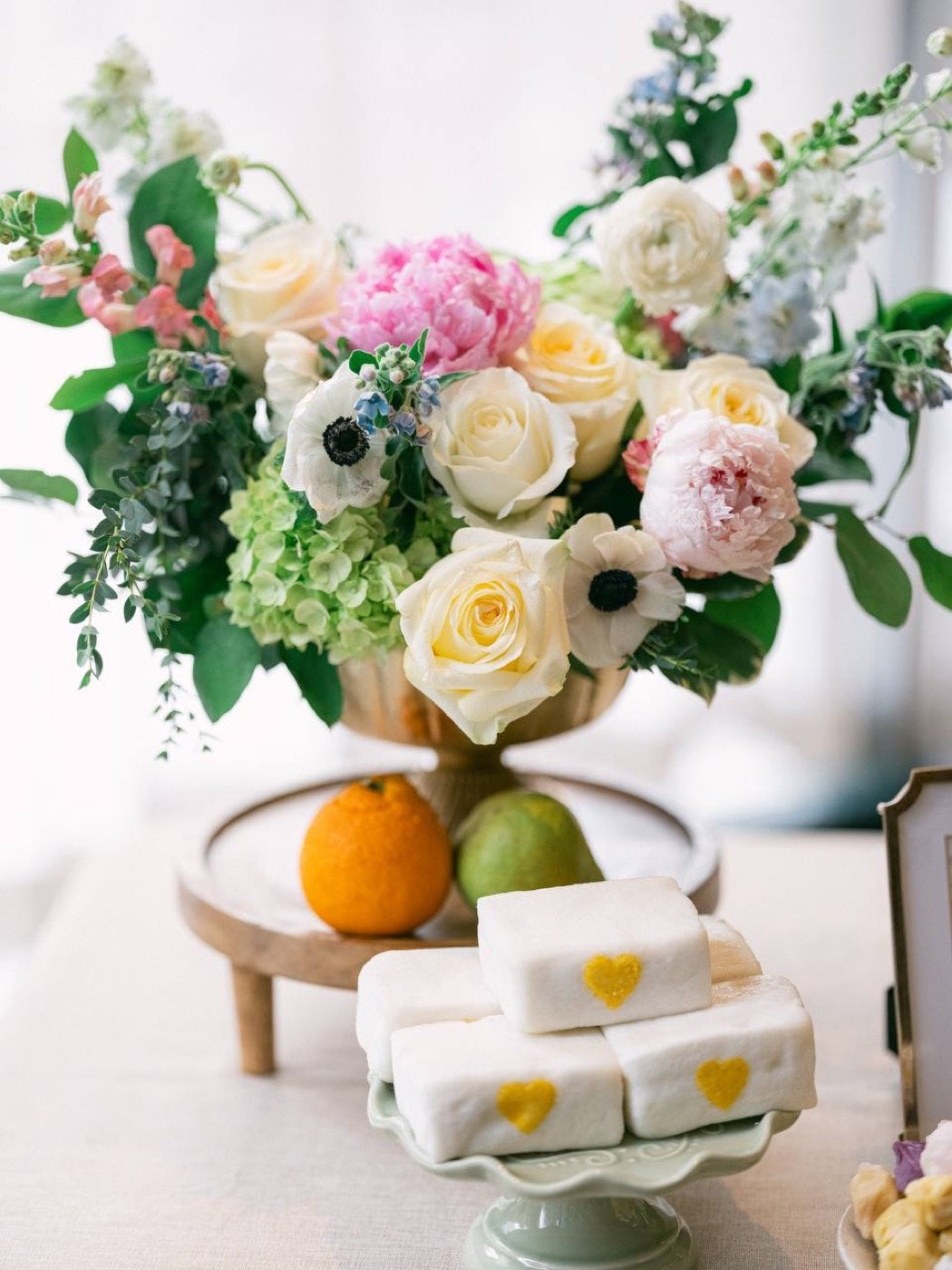 Dessert table styled by Novapetal Flower Lab, with a footed urn of white roses, peonies and anemones at one end and a low bowl arrangement at the other, beside a cake and stands of sweets