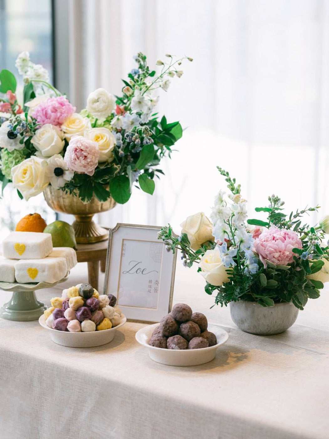 Close-up of a footed arrangement by Novapetal Flower Lab with white roses, a pink peony, anemones, snapdragon and green hydrangea, on a dessert table with fruit and petits fours