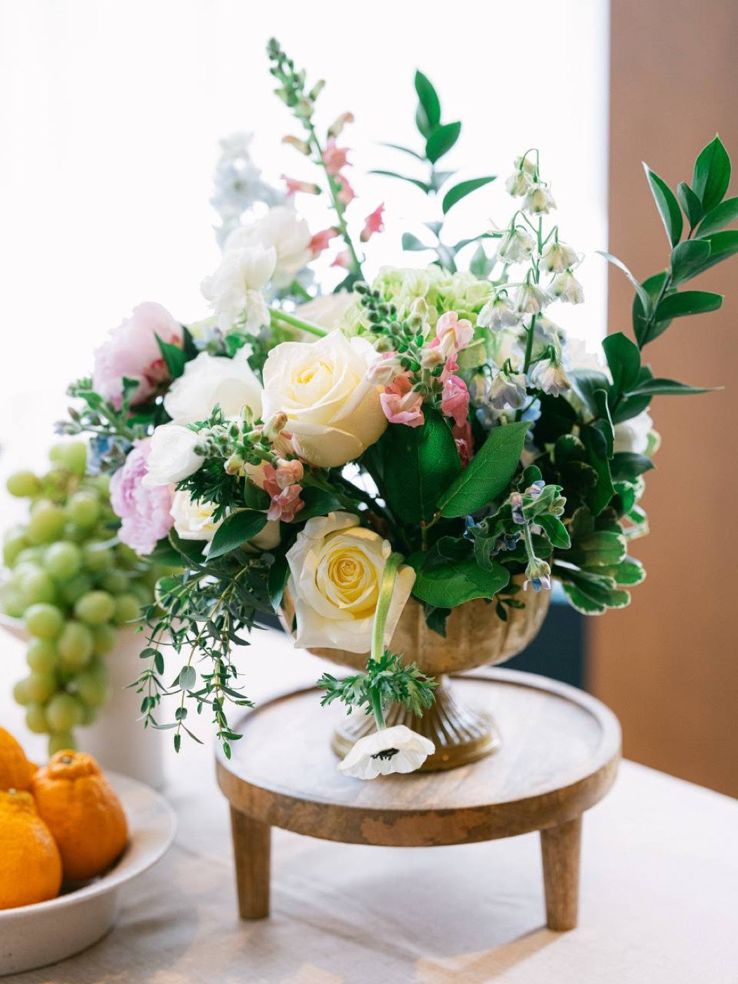 Footed brass urn arrangement by Novapetal Flower Lab with white roses, blush peony, anemones, snapdragon and delphinium, styled on a wooden riser beside grapes and citrus
