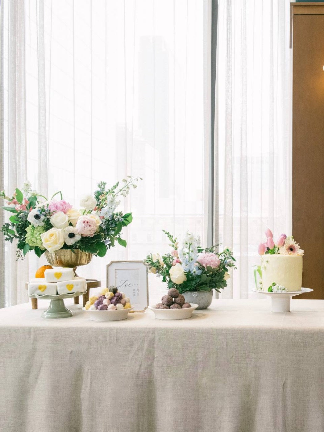 Event table by Novapetal Flower Lab with a tall urn arrangement, a low bowl of white roses and pink peonies and a framed name card, set with chocolates and fruit