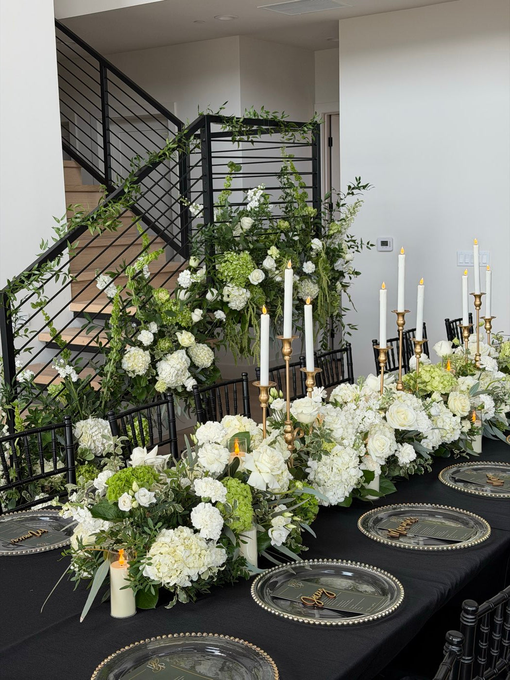 Long banquet table styled by Novapetal Flower Lab with white roses, hydrangea and green foliage, gold taper candles and a trailing greenery installation