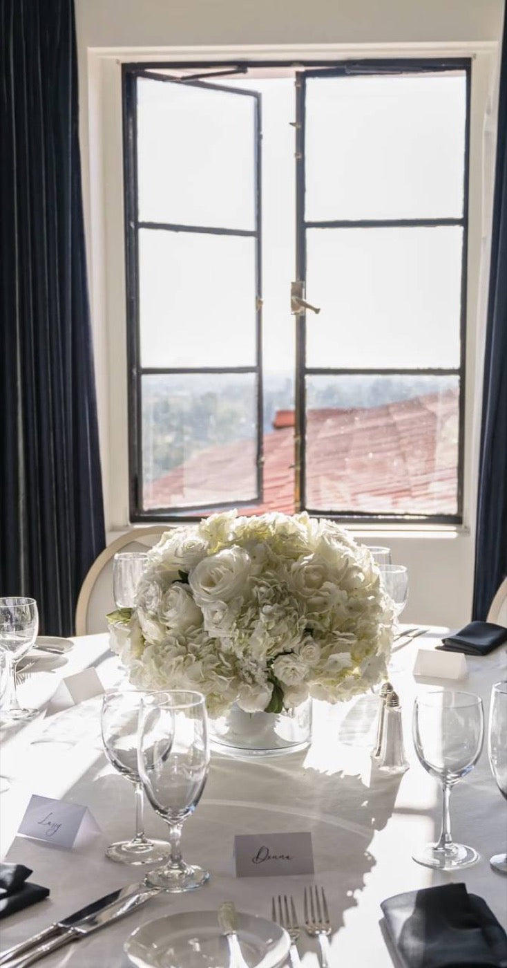 Low all-white centerpiece of roses, hydrangea and ranunculus by Novapetal Flower Lab on a round table set with place cards