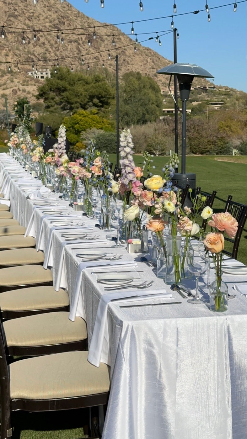Outdoor wedding table styled by Novapetal Flower Lab at an Arizona venue, with bud vases of peach garden roses, delphinium and ranunculus under string lights and desert mountains behind