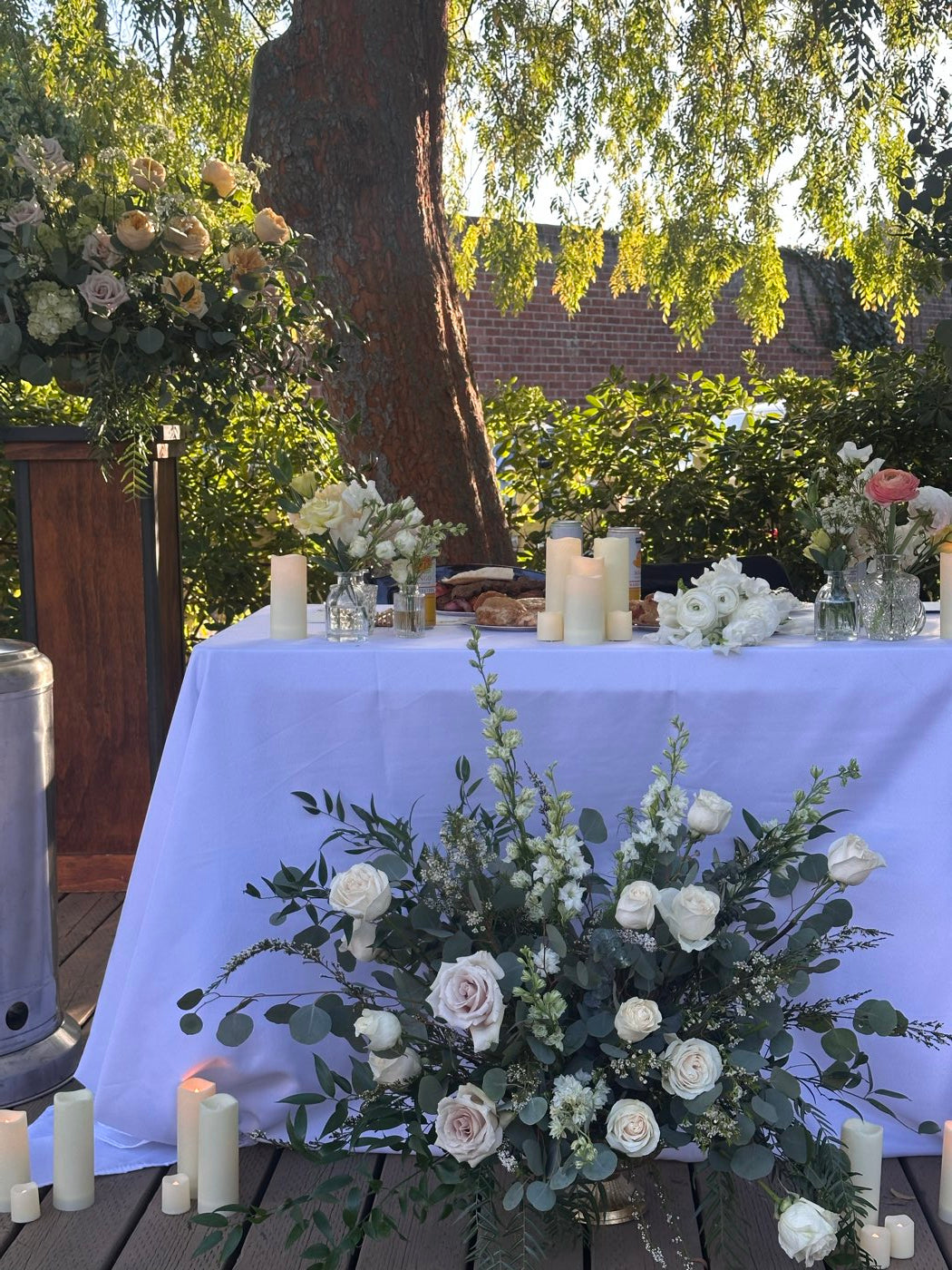Outdoor reception table by Novapetal Flower Lab with a blush and white rose arrangement in eucalyptus, pillar candles and bud vases under a tree