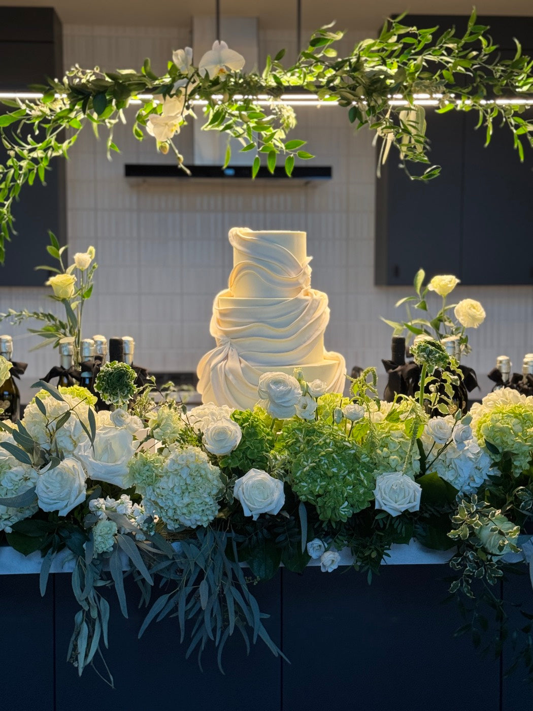 Wedding cake table by Novapetal Flower Lab banked with white roses, hydrangea and green viburnum, under a hanging greenery and orchid installation