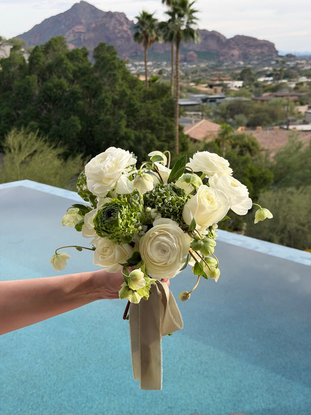 Bridal bouquet of white ranunculus, garden roses and hellebore by Novapetal Flower Lab, held above a pool with the Phoenix mountains and palm trees behind