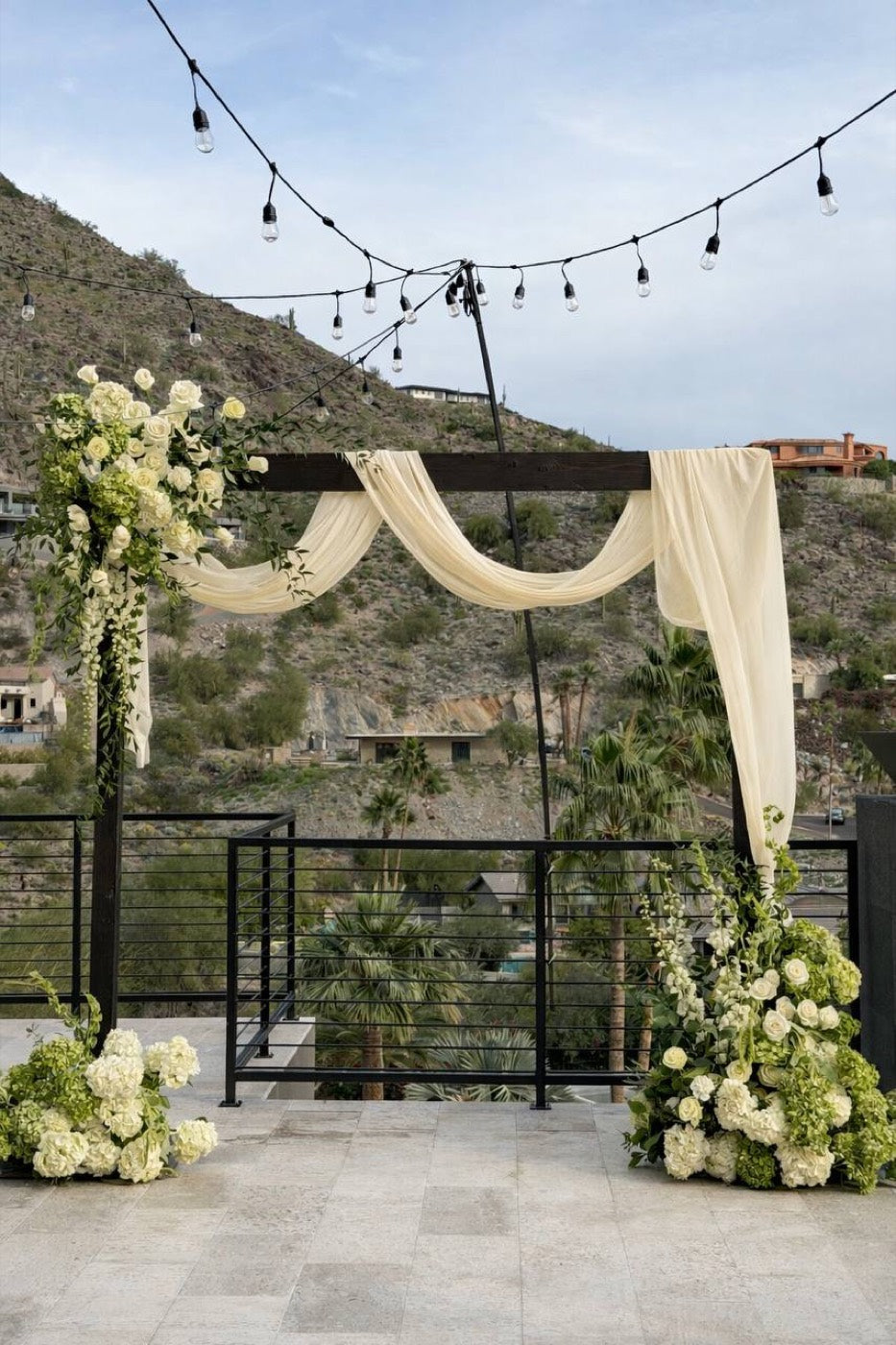 Ceremony arch by Novapetal Flower Lab on a Phoenix terrace, draped in ivory fabric with white roses, hydrangea and trailing greenery, the mountains and string lights behind