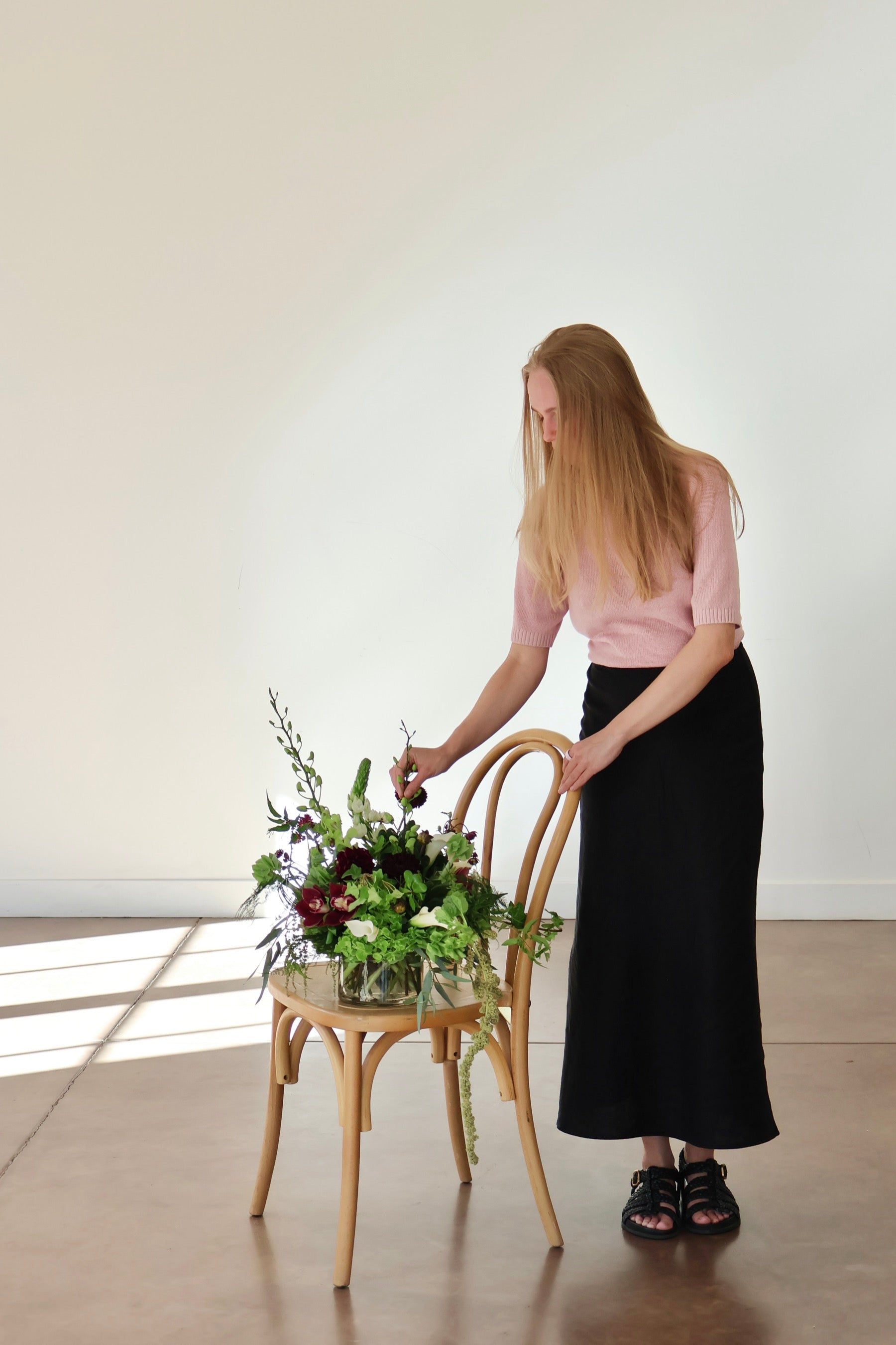 Novapetal Flower Lab designer finishing a burgundy and green arrangement on a bentwood chair