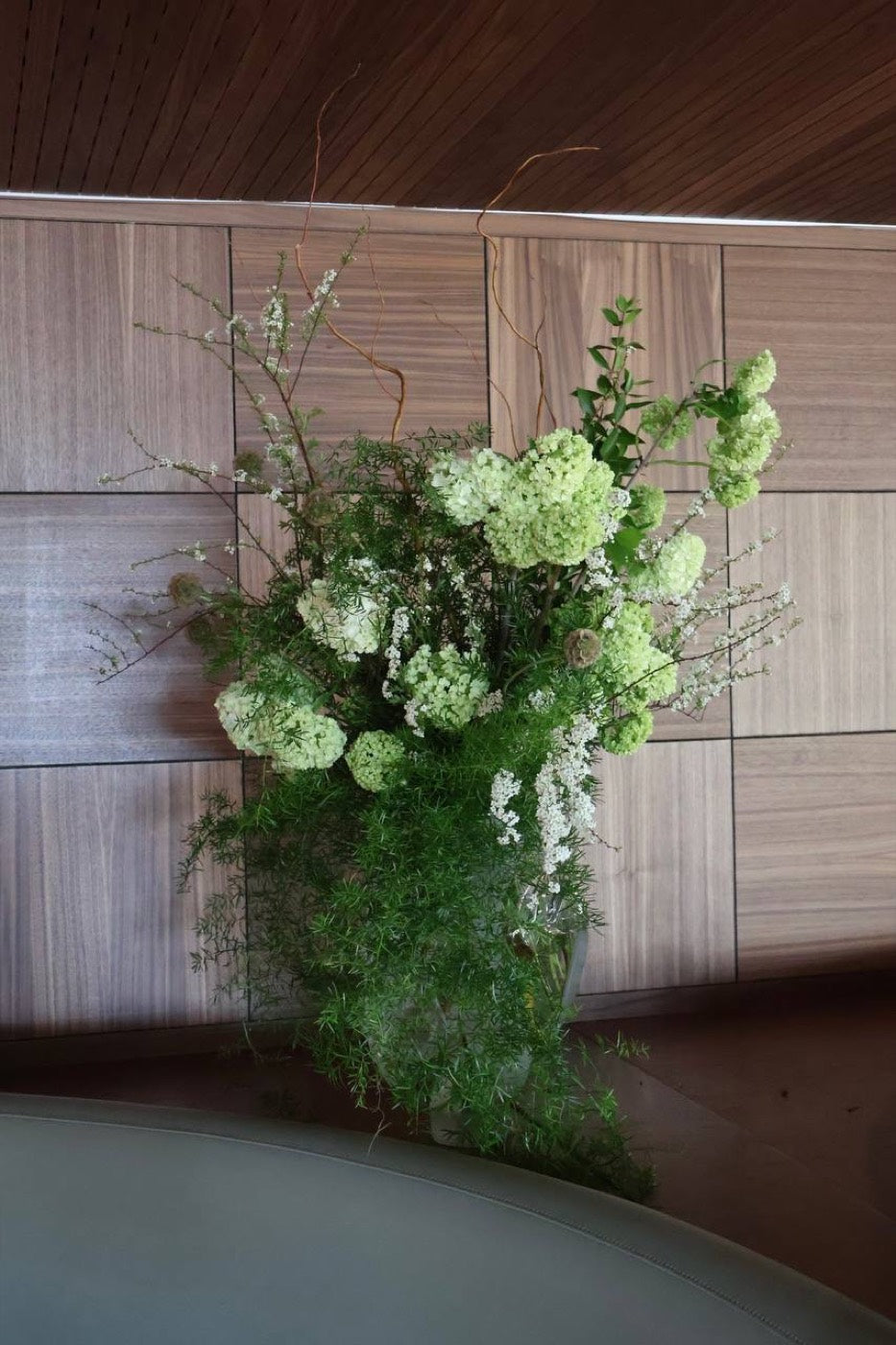 Green and white viburnum and spirea arrangement by Novapetal Flower Lab trailing over the edge of a restaurant table, set against wood panelling