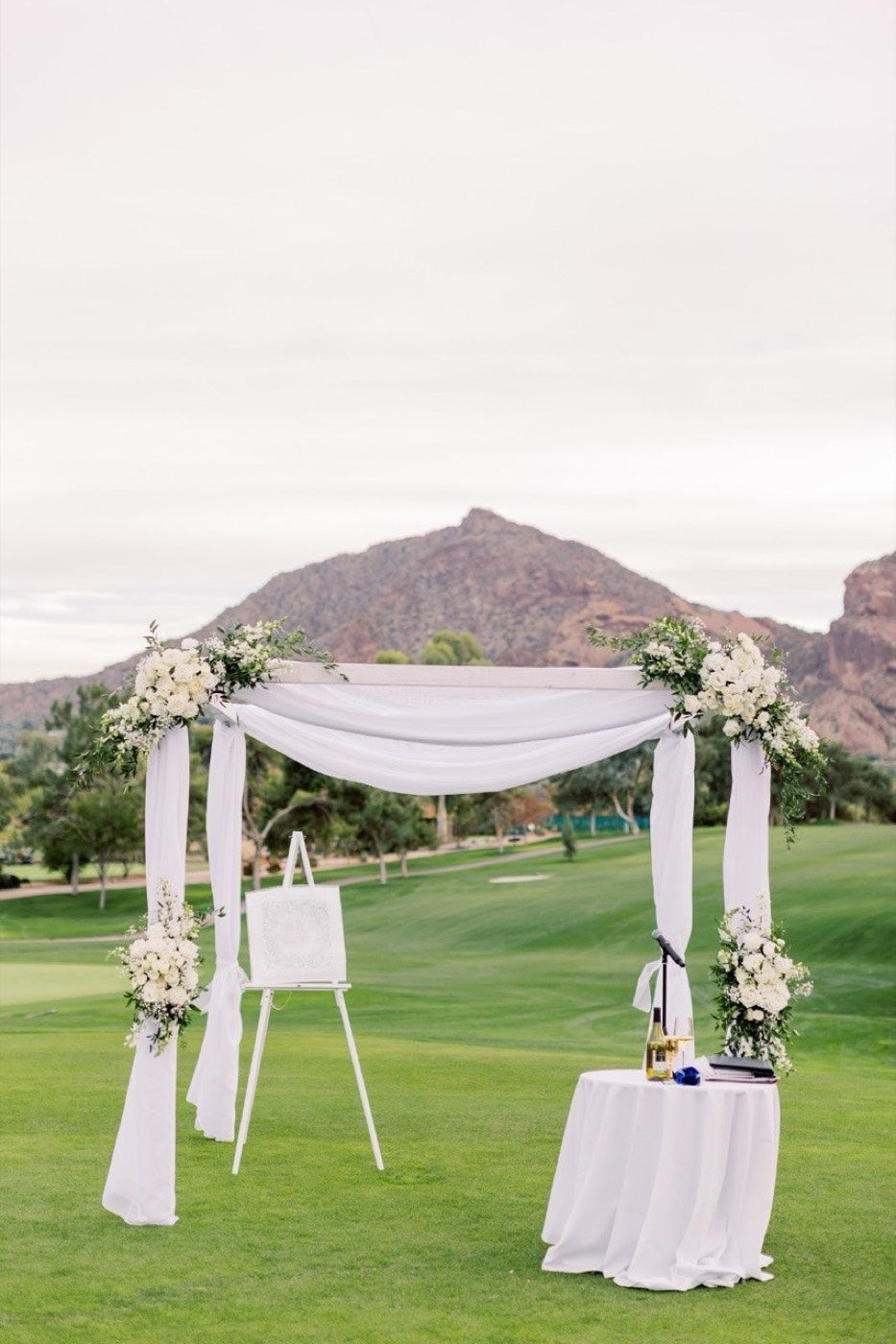 White draped ceremony arch by Novapetal Flower Lab on a golf course in Paradise Valley, dressed with white hydrangea and roses, Camelback Mountain behind