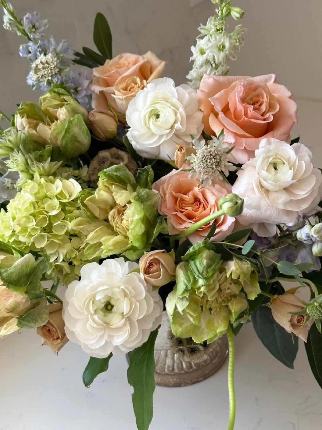 Peach and green bridal arrangement by Novapetal Flower Lab with garden roses, ranunculus, hellebore and hydrangea in a stone urn