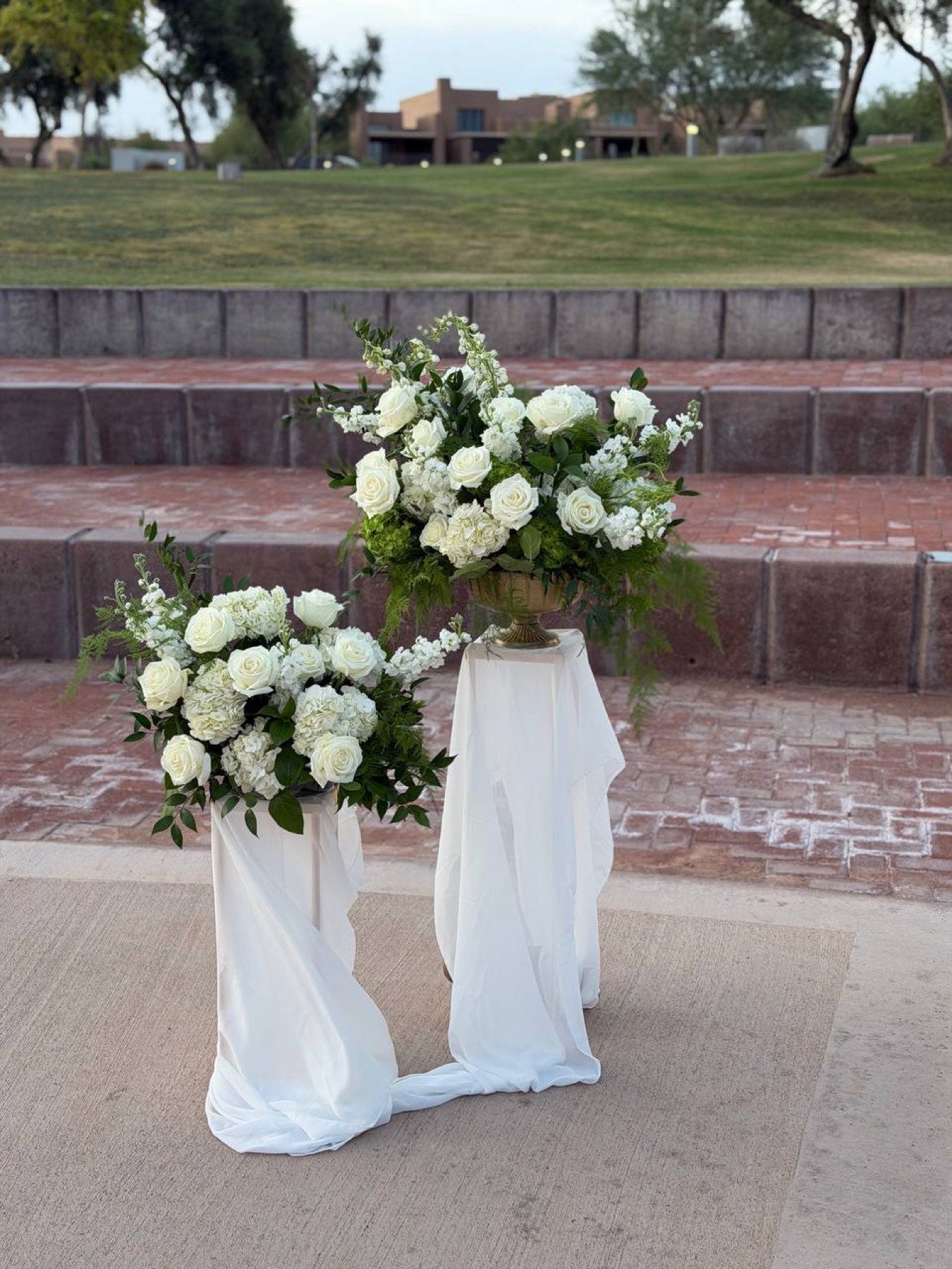 Two draped pedestals by Novapetal with white roses, hydrangea and stock, set on the brick steps of the ceremony lawn