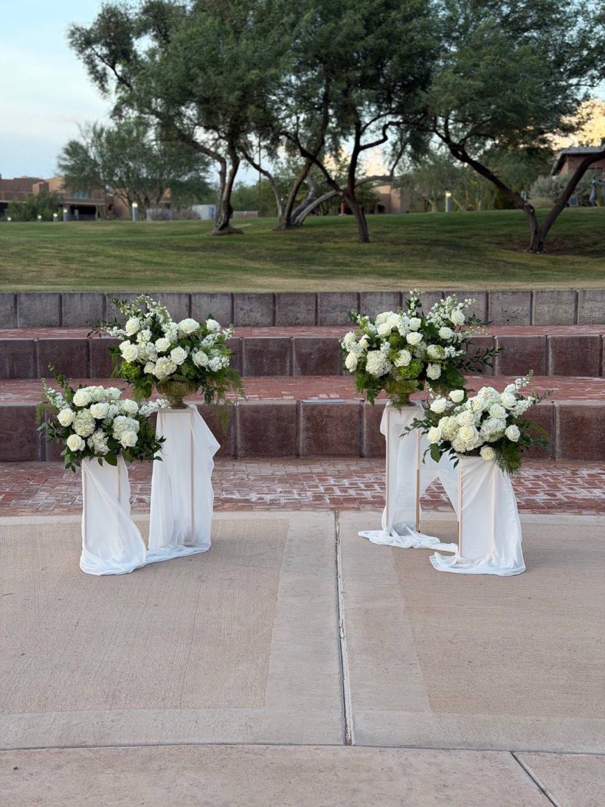 Four draped pedestals by Novapetal flanking the aisle, white roses and hydrangea against the desert trees and lawn
