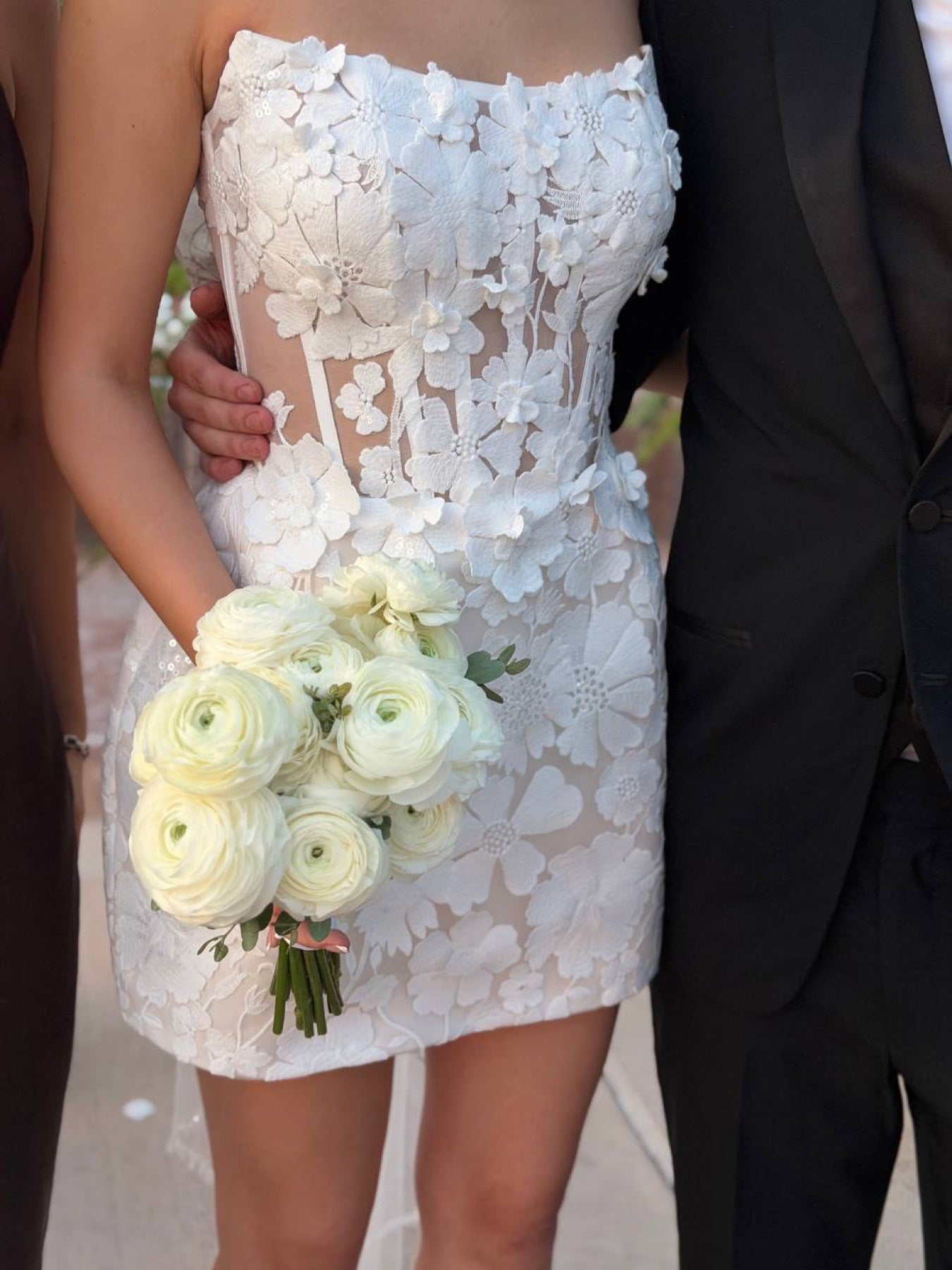 Bride in a short lace dress holding a white ranunculus bouquet by Novapetal, standing with the groom